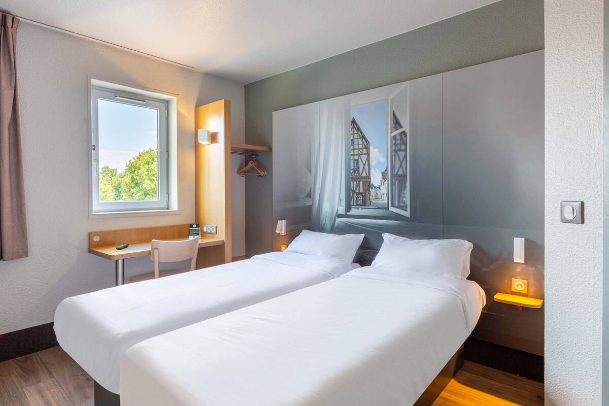 A hotel room with two beds, a desk, a chair, a window, and a wall mural.