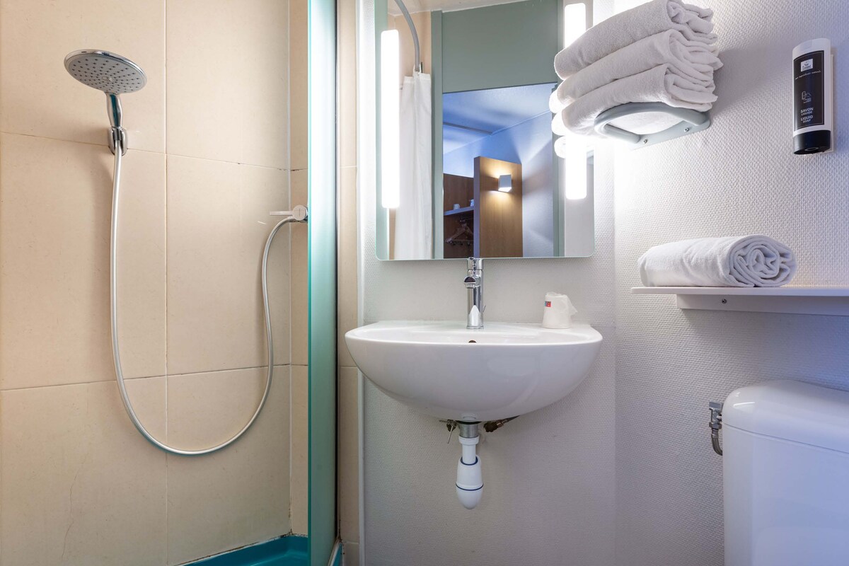 A bathroom with a shower, sink, mirror, towel rack, and a wall-mounted device.