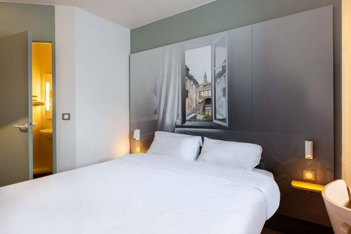 A hotel room with a bed, a painting of a street scene on the wall, and bedside lamps.