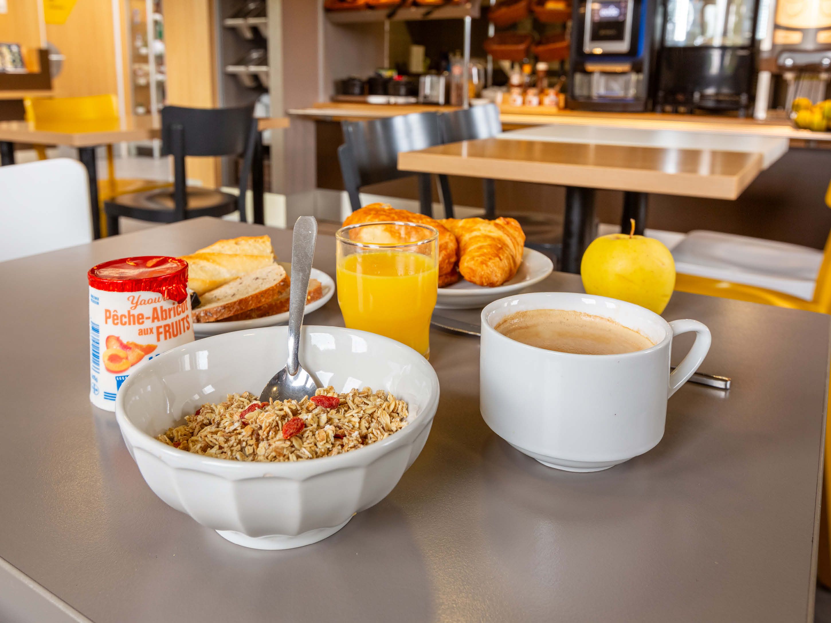 Un desayuno con un tazón de cereal, un vaso de jugo de naranja, un croissant, una taza de café y un yogurt de frutas en lata.