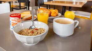A breakfast setting with a bowl of cereal, a glass of orange juice, a croissant, a coffee cup, and a can of fruit yogurt. - B&B Hotel Chartres 2 Le Coudray (Le Coudray)