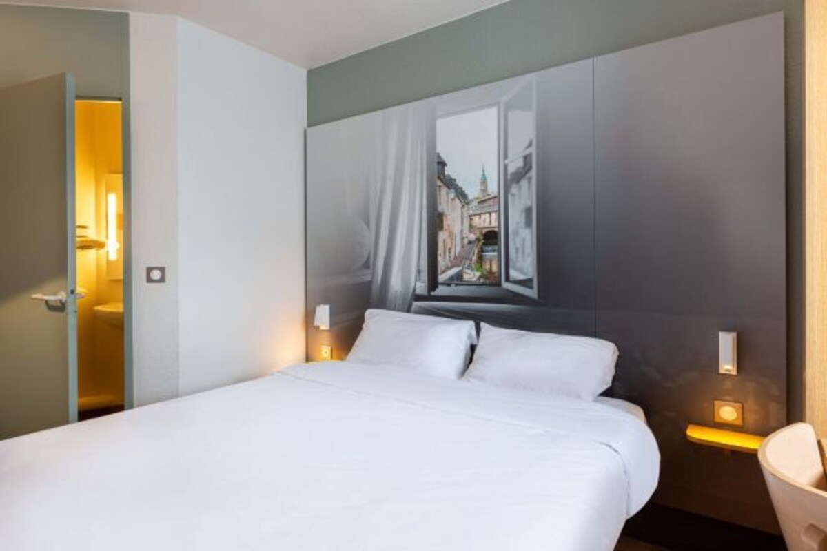 A hotel room with a bed, a painting of a street scene on the wall, and bedside lamps.