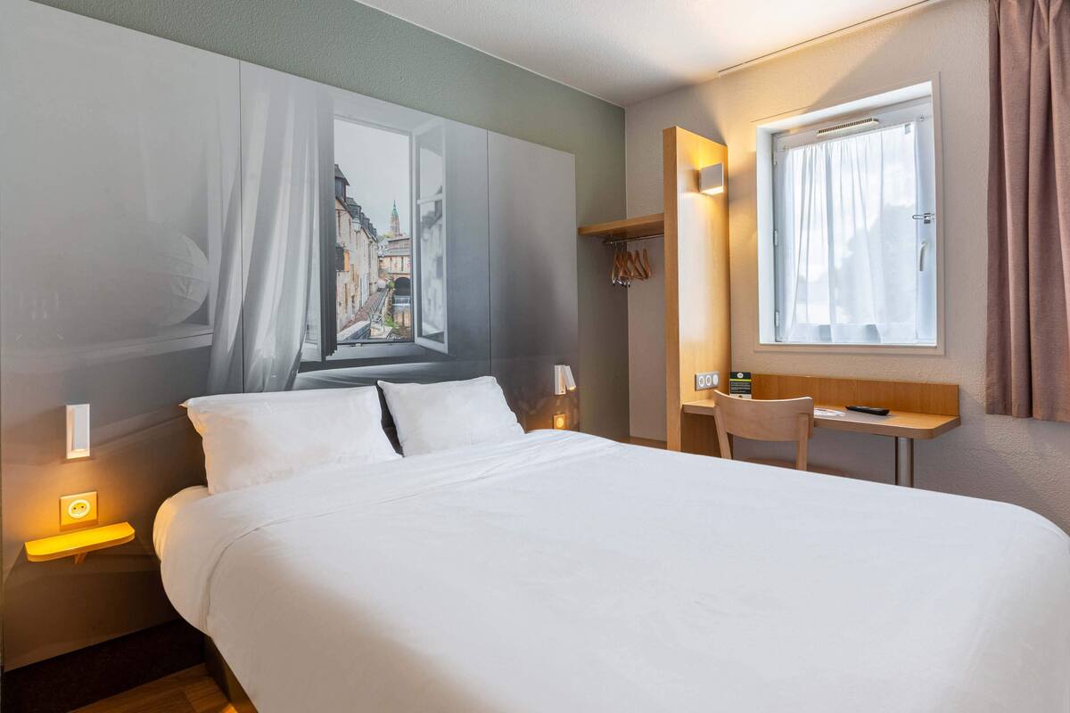 A hotel room with a bed, bedside table, lamp, window, and a small desk with a chair.