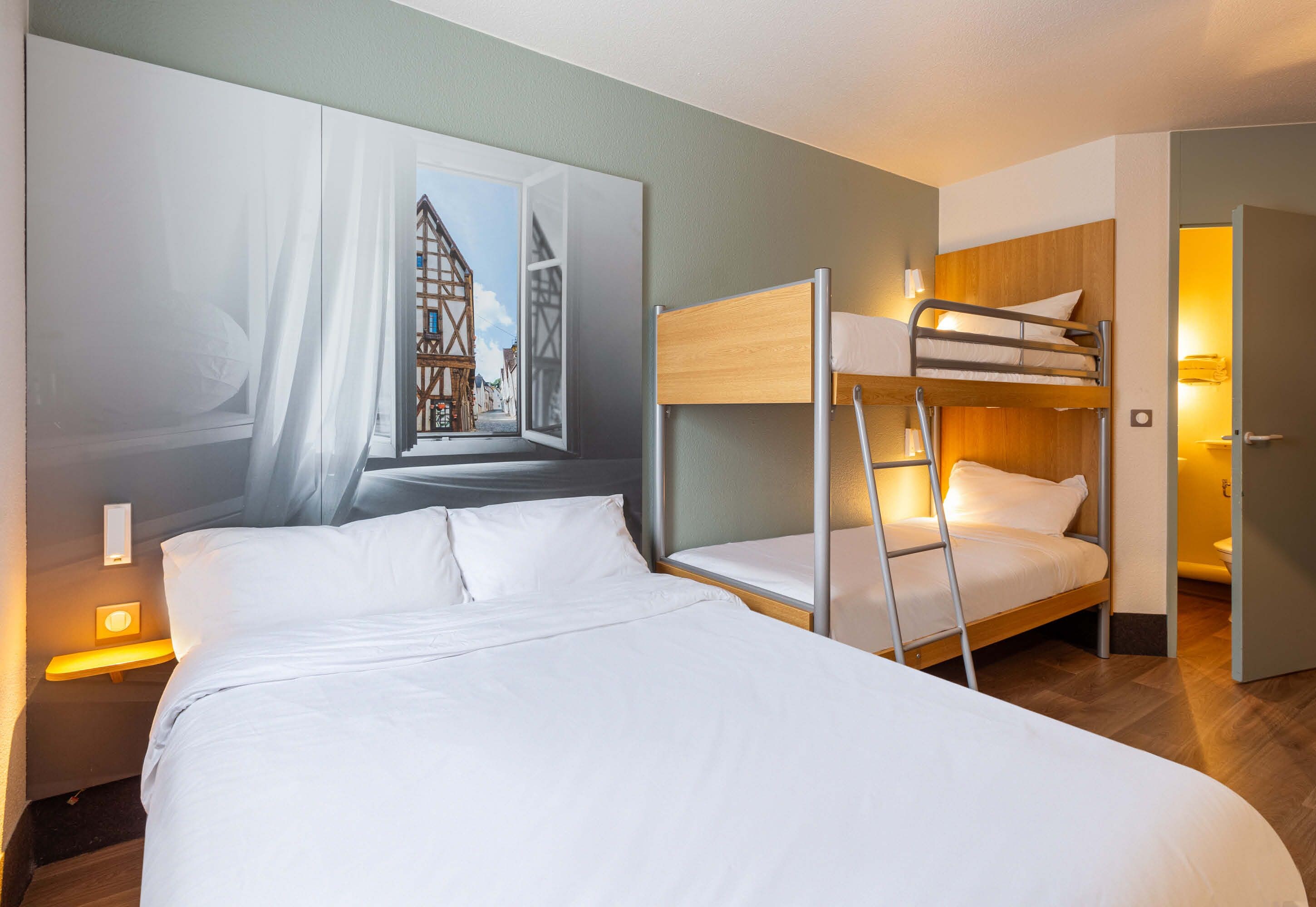 A hotel room with a bunk bed, a large window with a view of a building, and a bedside lamp.