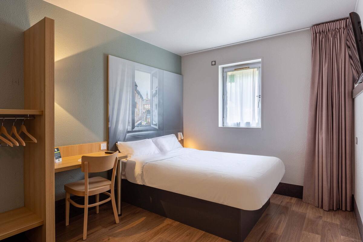A compact hotel room with a single bed, a desk, a chair, a wardrobe, and a window with curtains.