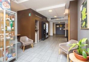 A hotel lobby with seating, a glass display case, a refrigerator, and a potted plant. - B&B Hotel Chartres 2 Le Coudray (Le Coudray)