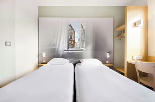 Habitación de hotel con dos camas individuales, un escritorio, una silla y vistas a un edificio tradicional a través de un amplio ventanal.