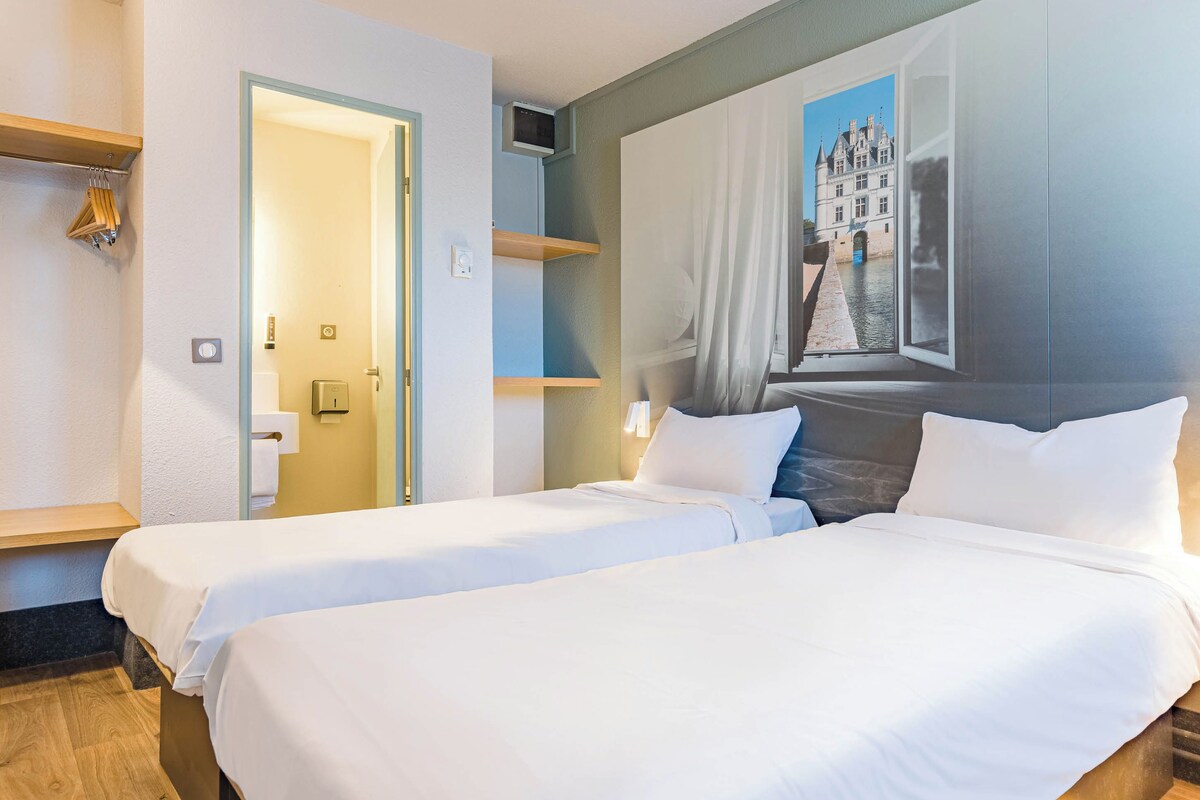 A hotel room with two beds, a painting of a building, a wooden shelf, and a door leading to another room.