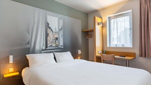 A hotel room with a bed, bedside table, lamp, window, and a small desk with a chair. - B&B Hotel Chartres 2 Le Coudray (Le Coudray)