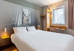 Ett hotellrum med en säng, ett nattduksbord, en lampa, ett fönster och ett litet skrivbord med en stol.