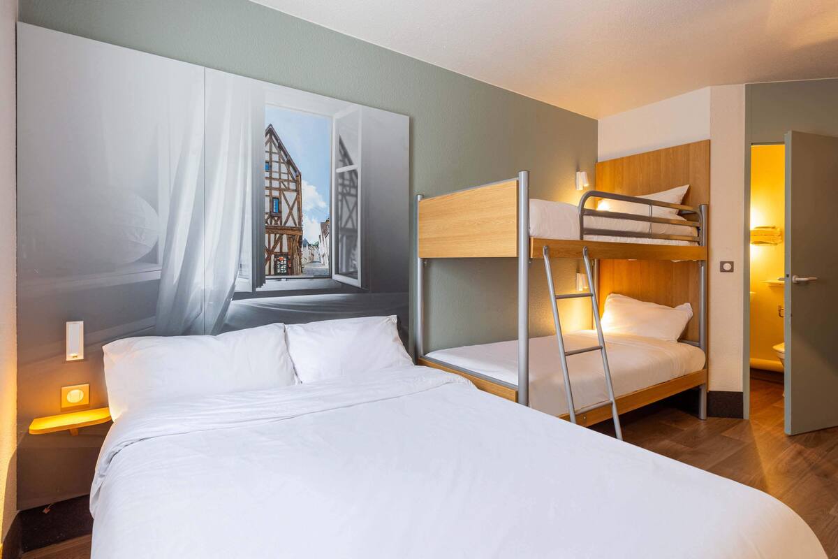 A hotel room with a bunk bed, a large window with a view of a building, and a bedside lamp.