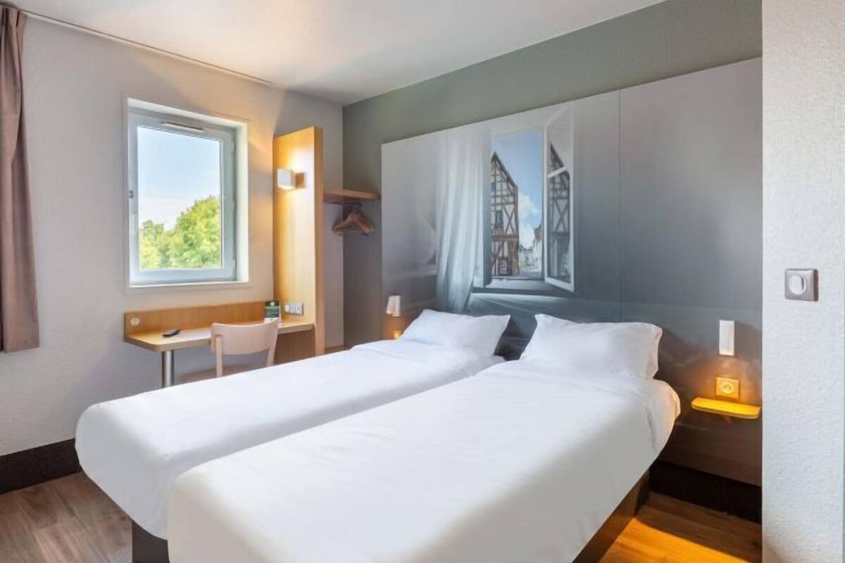 A hotel room with two beds, a desk, a chair, and a window with a view of greenery.