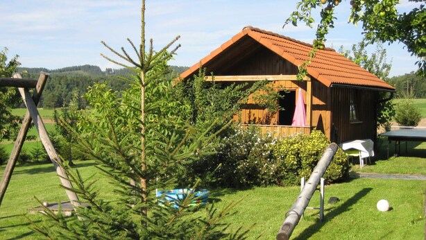 Eine Holzhütte mit geneigtem Dach, umgeben von üppigem Grün und blauem Himmel.
