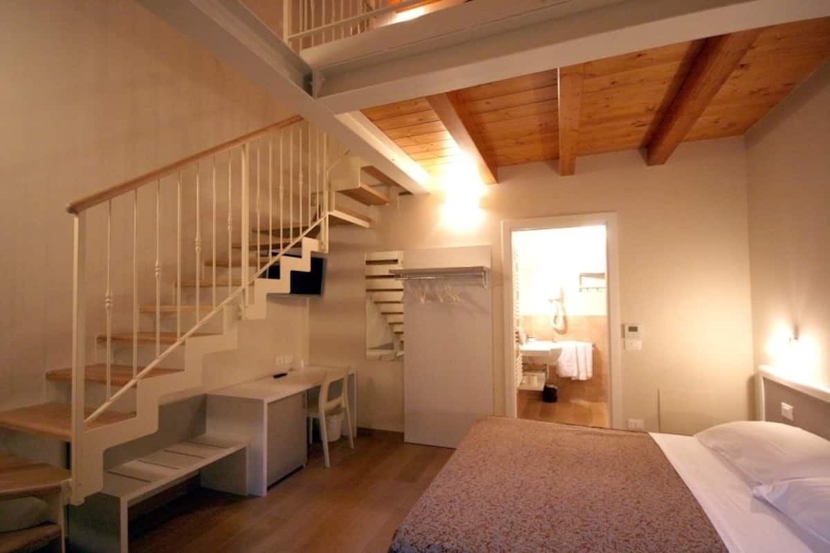 A bedroom with a wooden ceiling, a staircase, a bed, a desk, and a bathroom visible through an open door.