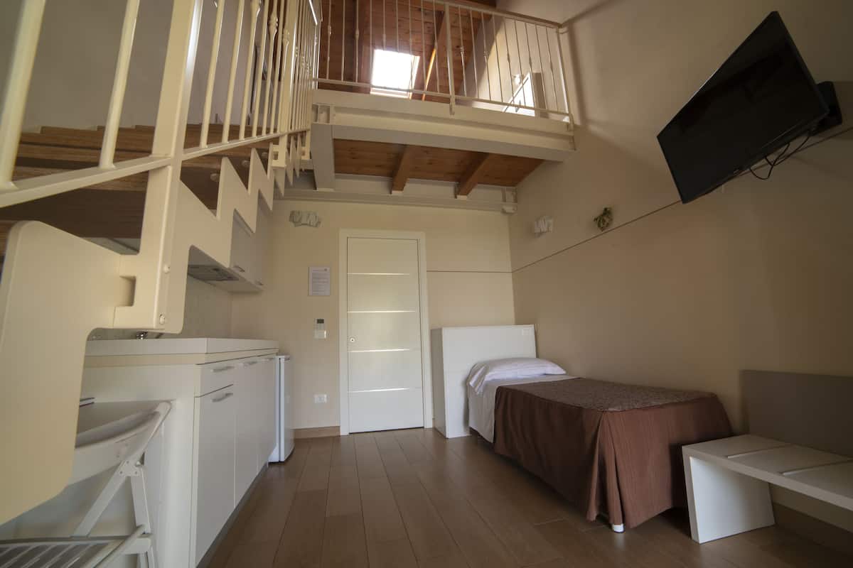 A compact hotel room with a kitchenette, a single bed, a small table, and a TV mounted on the wall.