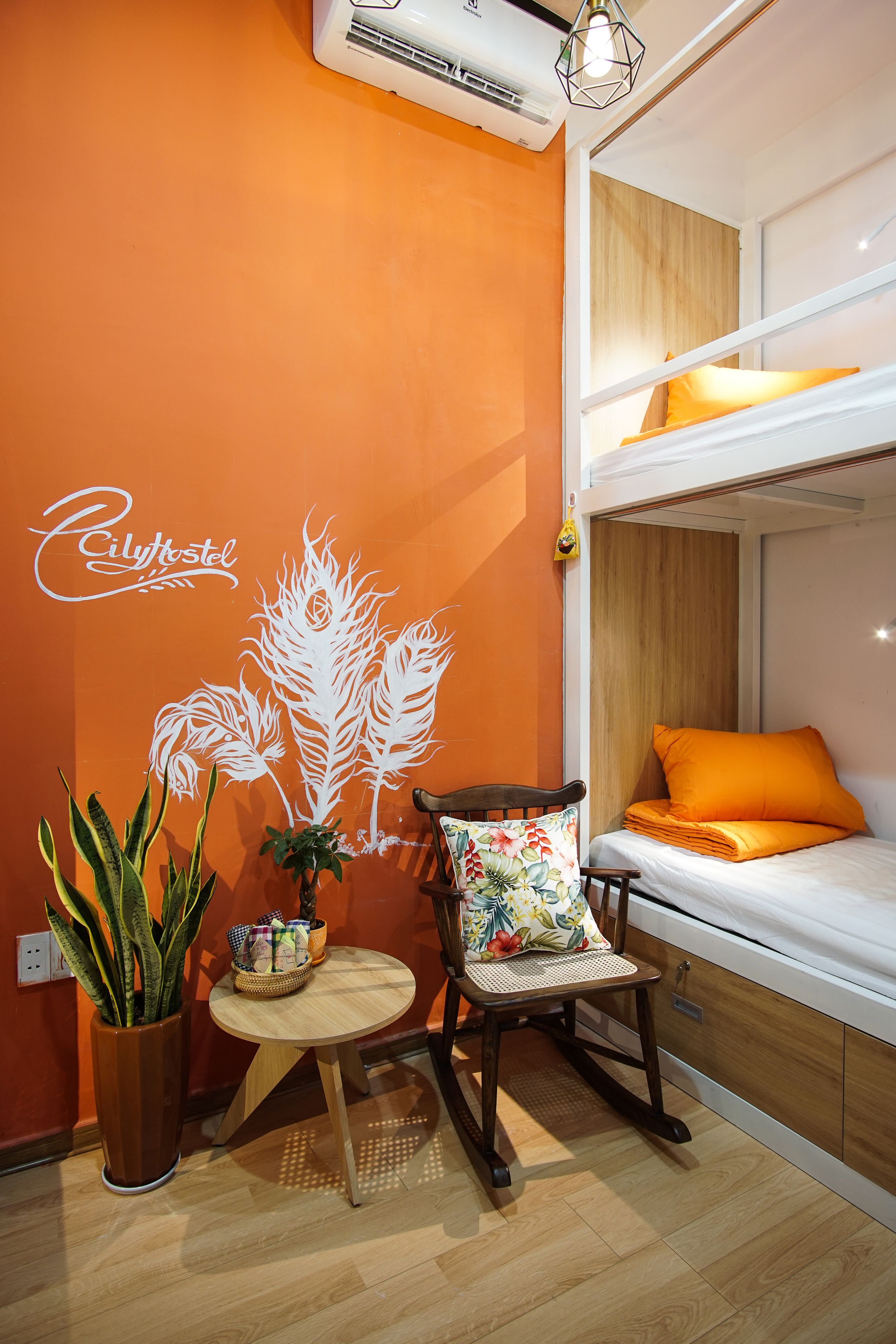 Una habitación con literas, una silla con cojín floral, una mesita con una cesta y una planta en maceta.