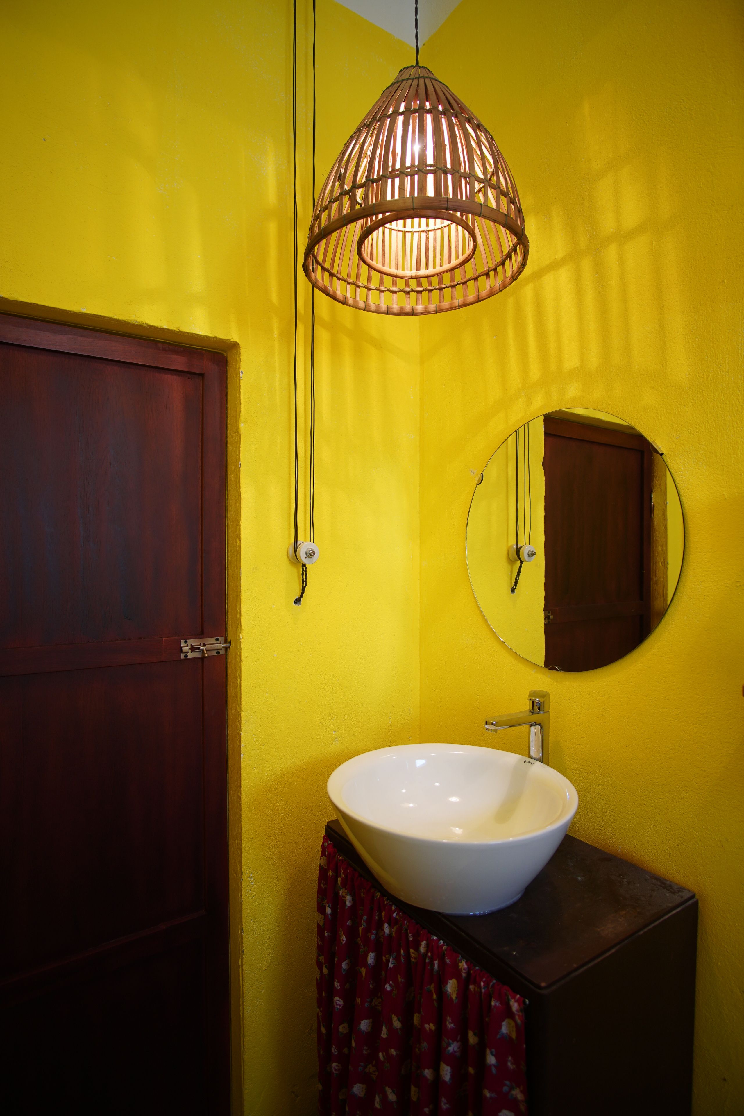 Een badkamer met een gele wand, een houten deur, een ronde spiegel, een wit wastafel en een hanglamp.