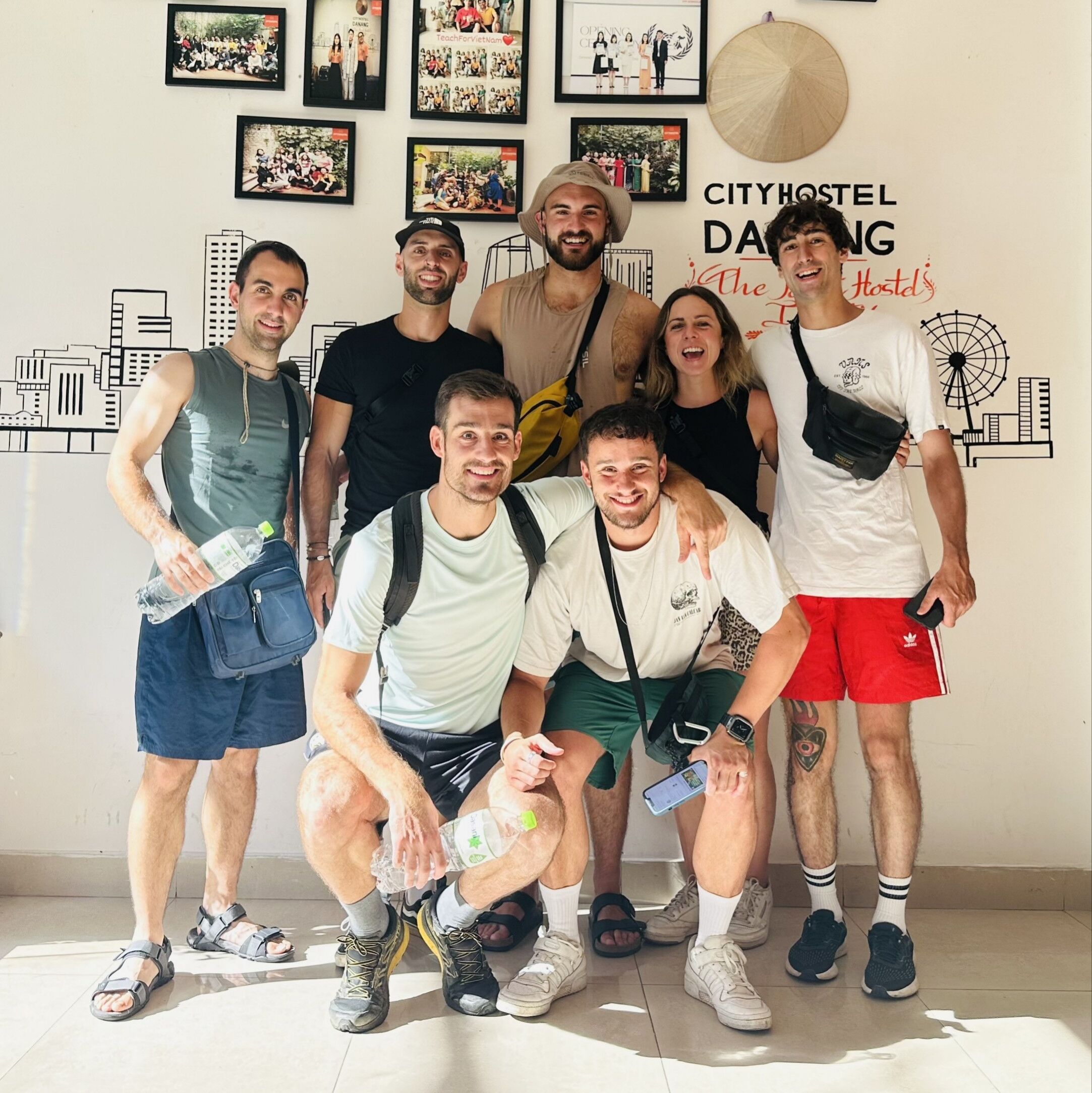 Un grupo de personas posando frente a una pared con fotos enmarcadas y un letrero que dice "CITYHOSTEL DANG".
