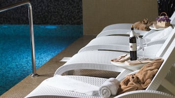 Un área de descanso junto a la piscina con sillones blancos acolchados, una botella de vino, copas y toallas.
