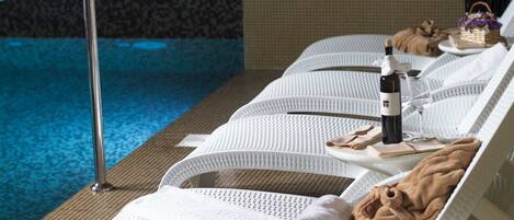 A poolside lounge area with white cushioned chairs, a bottle of wine, glasses, and towels.