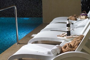 A poolside lounge area with white cushioned chairs, a bottle of wine, glasses, and towels.