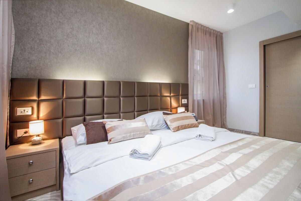 Habitación de hotel con una cama grande, una mesita de noche con lámpara, un enchufe en la pared y una puerta.
