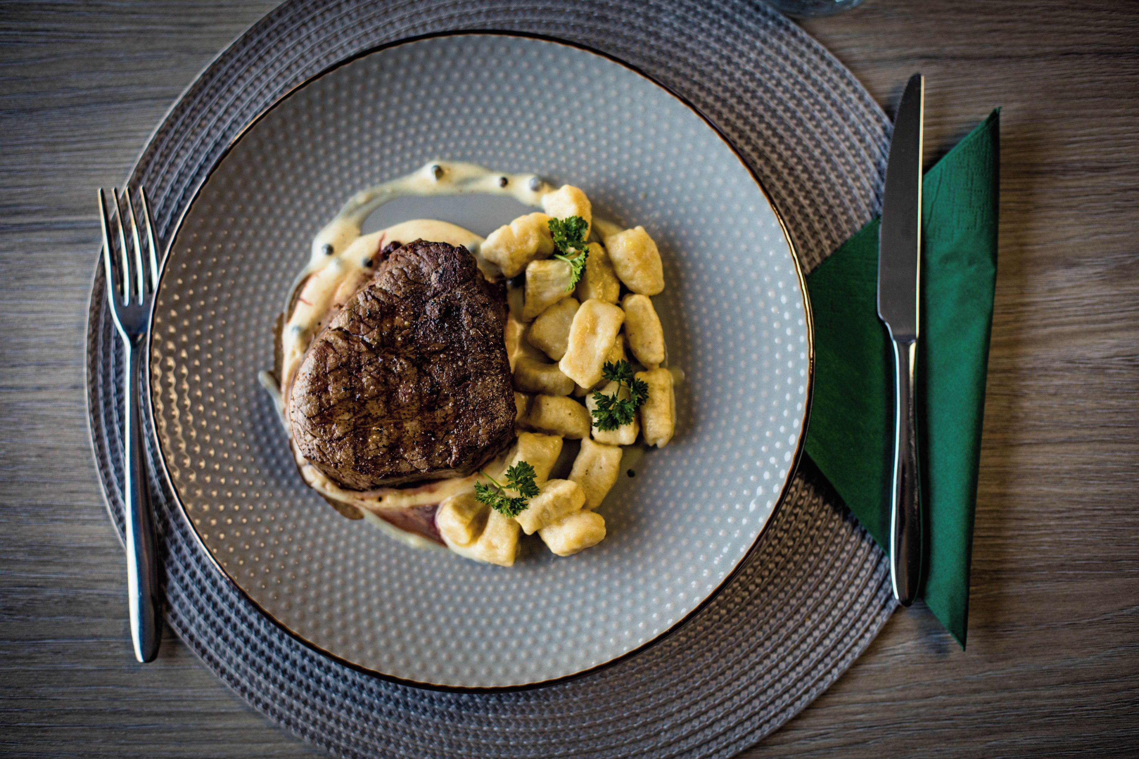 A plate with a steak, gnocchi, and sauce on a textured plate, set on a wooden table with a fork and knife.