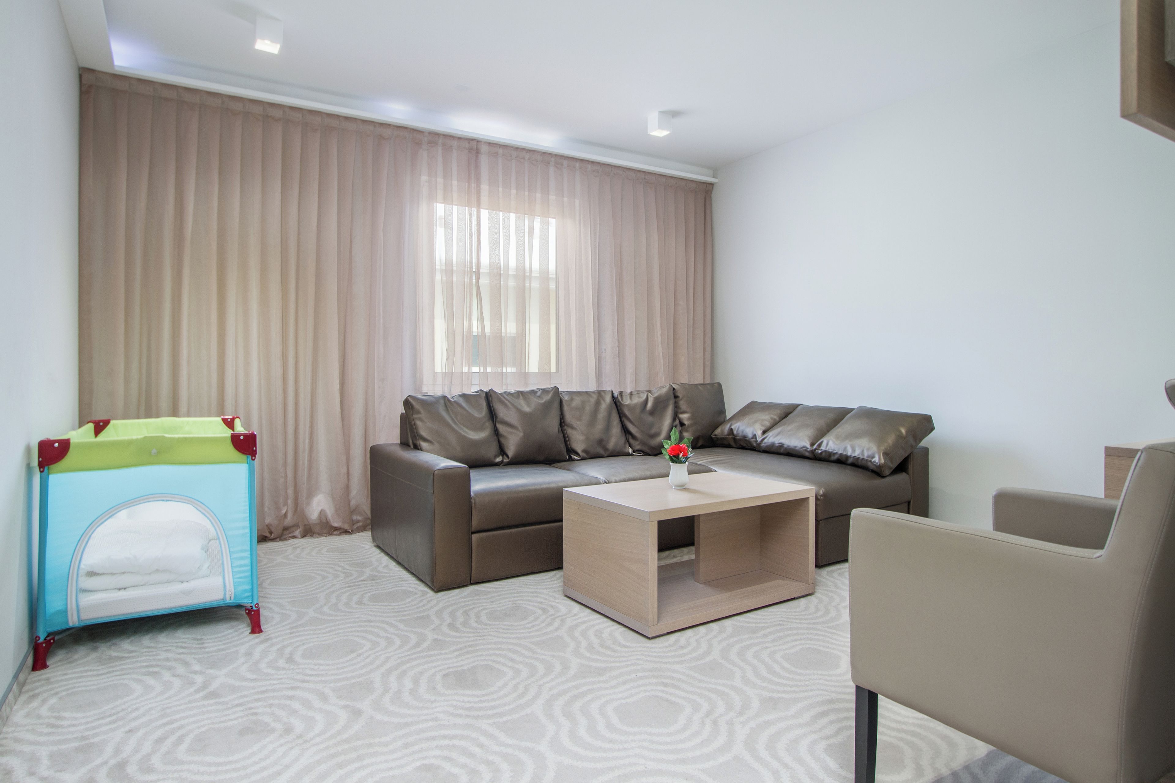 A modern living room with a brown leather sofa, a small coffee table, a beige armchair, and a child’s playpen.