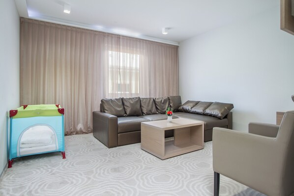 A modern living room with a brown leather sofa, a small coffee table, a beige armchair, and a child’s playpen. - Royal Airport (Velika Gorica)
