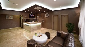 Zona d'entrada d'hotel amb taulell de recepció, un rellotge, un sofà i una planta en test.
