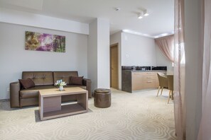 A modern living room with a brown leather sofa, a wooden coffee table, and a small ottoman. A painting is on the wall, and a kitchen area is visible in the background.