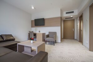 A modern hotel room with a sofa, a small table with a vase, a chair, a television mounted on the wall, and a door.