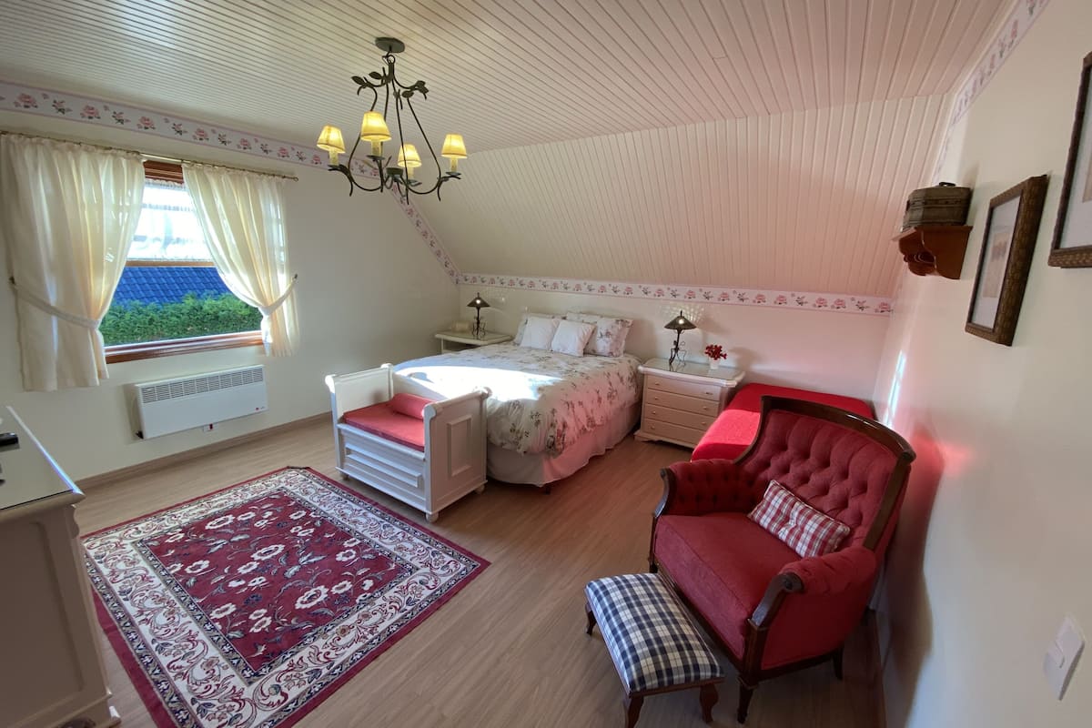 A cozy bedroom with a bed, a red armchair, a small table, a nightstand, a lamp, a chandelier, a window with curtains, and a radiator.