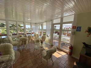 A sunlit room with white chairs and a glass table, large windows offering a view of the outdoors, and a fire extinguisher on the wall. - Pousada Chateau Colinas (Campos do Jordão)