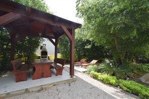 A covered outdoor seating area with benches and a table, surrounded by greenery and a building with a chimney. - Studio with air conditioning (Malinska)