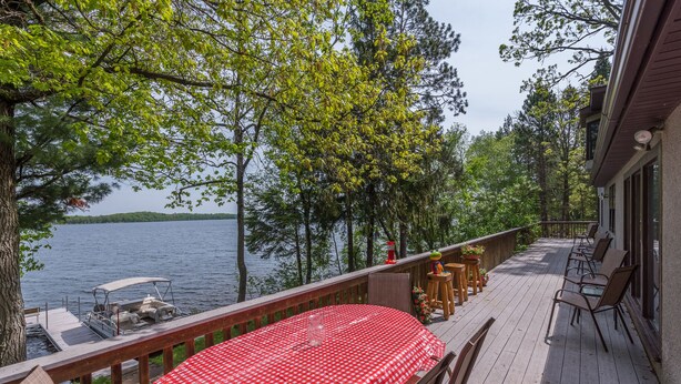 A wooden deck with a red and white checkered table and chairs, overlooking a lake with boats and trees.
