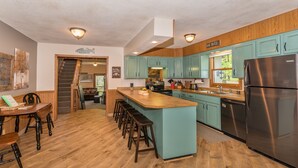 A kitchen with teal cabinets, a central island with stools, a stainless steel refrigerator, and a wooden staircase leading to another room.
