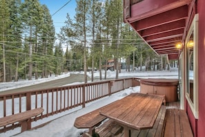 Eine Holzterrasse mit Whirlpool und Holztisch, umgeben von einem Geländer, mit Blick auf eine verschneite Landschaft und Bäume.