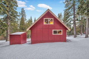 Eine rote Holzhütte mit zwei Fenstern, umgeben von Bäumen und Schnee.
