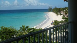 A beach view from a balcony with a railing, clear turquoise water, and a sandy shore with palm trees and beach umbrellas. - On Dover Beach, close to golf, surfing and much more. Discounts available (St-Lawrence Gap)