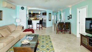 A living room with a sofa, coffee table, and TV stand. There is a dining area with a table and chairs. The room has a ceiling fan and a kitchen in the background. - On Dover Beach, close to golf, surfing and much more. Discounts available (St-Lawrence Gap)