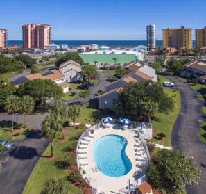 Un resort con una amplia piscina circular rodeada de sillones de descanso y sombrillas, ubicado en una zona residencial con edificios altos cercanos y una vista despejada del océano.