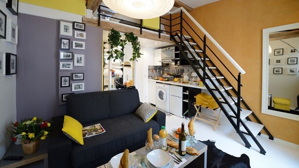 A modern living room with a black sofa, a dining table set with plates and glasses, a staircase, and a kitchen area visible through an open door. - D4DG Watergate (Paris)