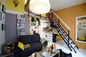 Un salon moderne avec un canapé noir, une table à manger dressée avec des assiettes et des verres, un escalier et un coin cuisine visible à travers une porte ouverte.