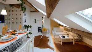 A modern loft apartment with a high ceiling, exposed wooden beams, a dining area with a white table and orange placemats, a living area with a white sofa and a brown leather rug, and a television mounted on the wall.