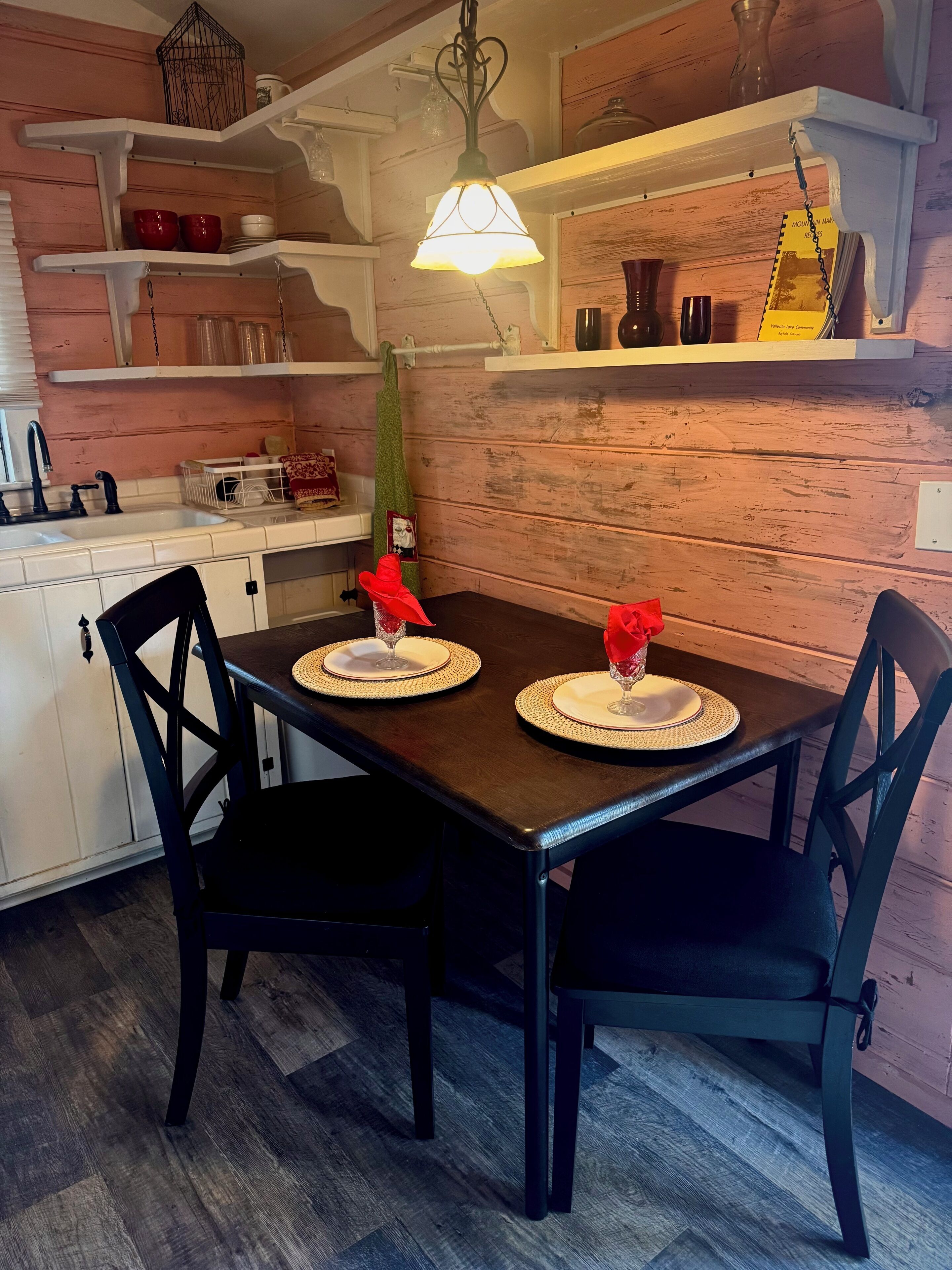 A dining area with a table set for two, chairs, a hanging light fixture, and shelves above the table.
