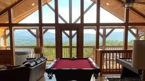 Un interior de madera con mesa de billar, un sofá y vistas a montañas a través de amplios ventanales.