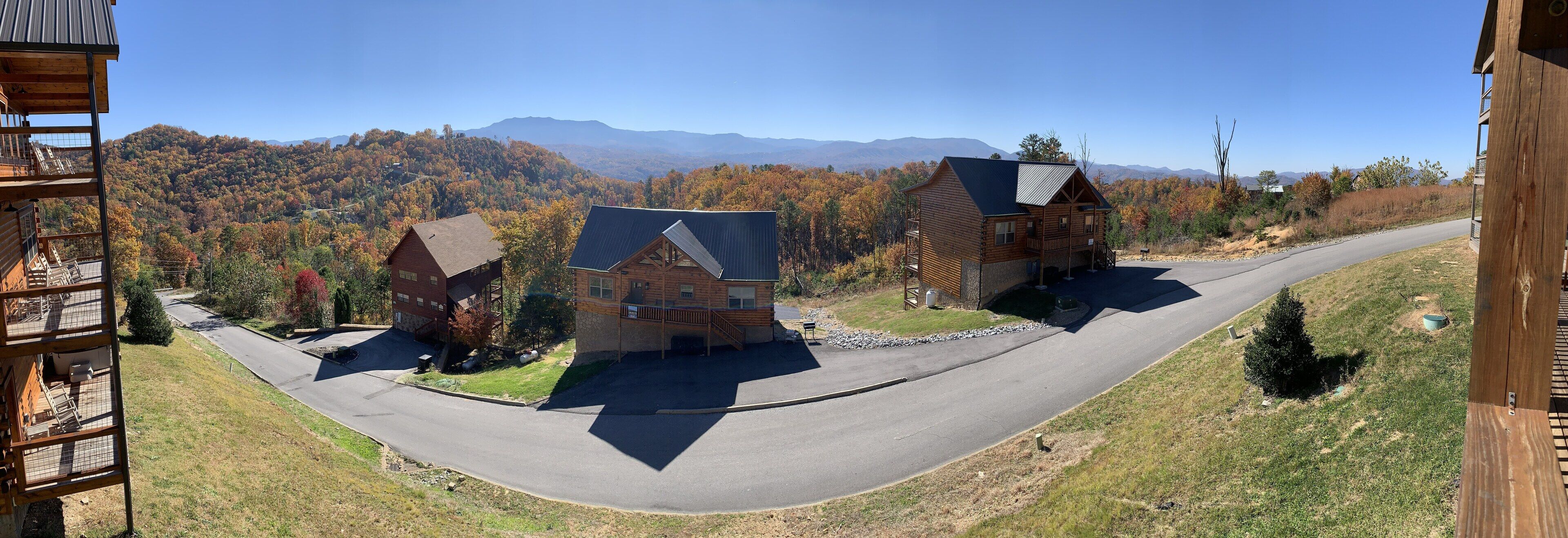 A view of a mountain cabin with a curved driveway, surrounded by autumn foliage and distant mountains.