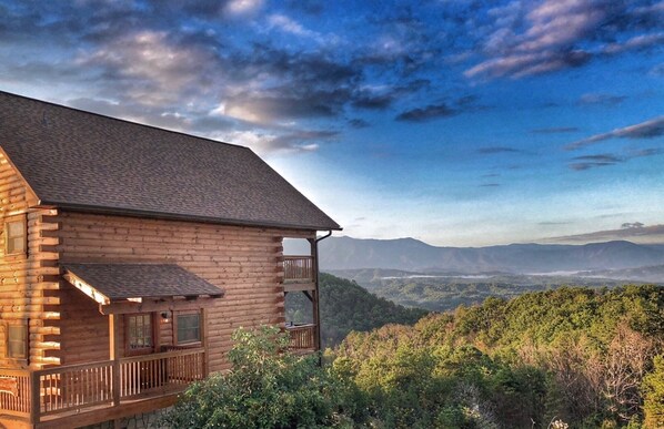 A log cabin with a balcony, surrounded by forested hills and mountains.