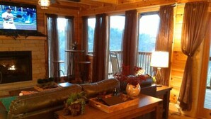 Una habitación acogedora estilo cabaña con chimenea, un televisor montado en la pared, una mesa de comedor de madera con un jarrón de flores y un amplio ventanal con cortinas.